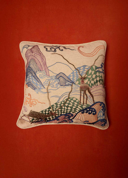 A decorative embroidered cushion featuring a landscape motif with mountains, trees, and a deer, crafted from fabric with colorful threads in shades of purple, green, blue, and red. The cushion has a neutral background and is set against a red surface.