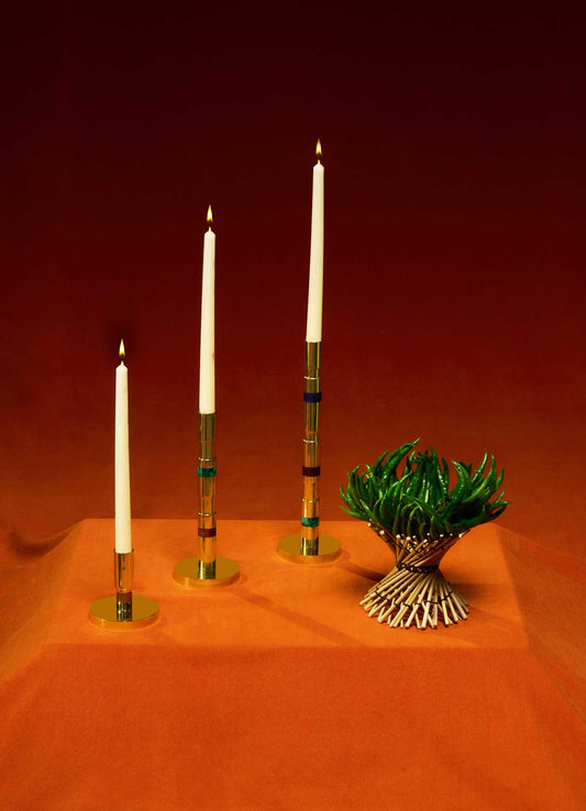 Three white taper candles in VIYA Liberty Candle Holders are arranged on an orange surface beside a small green plant, evoking Art Deco elegance against a striking red-orange gradient background.