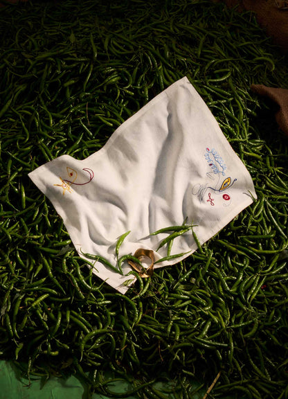 The Viya Evil Eye Hand Embroidered White Placemat (Set of 2) features abstract Nazar motif faces on fine table linen, styled here atop green chili peppers with a few chilies and a protective totem ring near the edge.