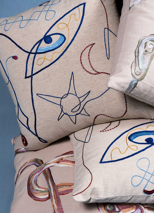 Viya’s Evil Eye Ecru Hand Embroidered Cushion Covers (18"x18") are stacked together, showcasing vibrant Nazar-inspired designs on a beige background against a light blue backdrop.