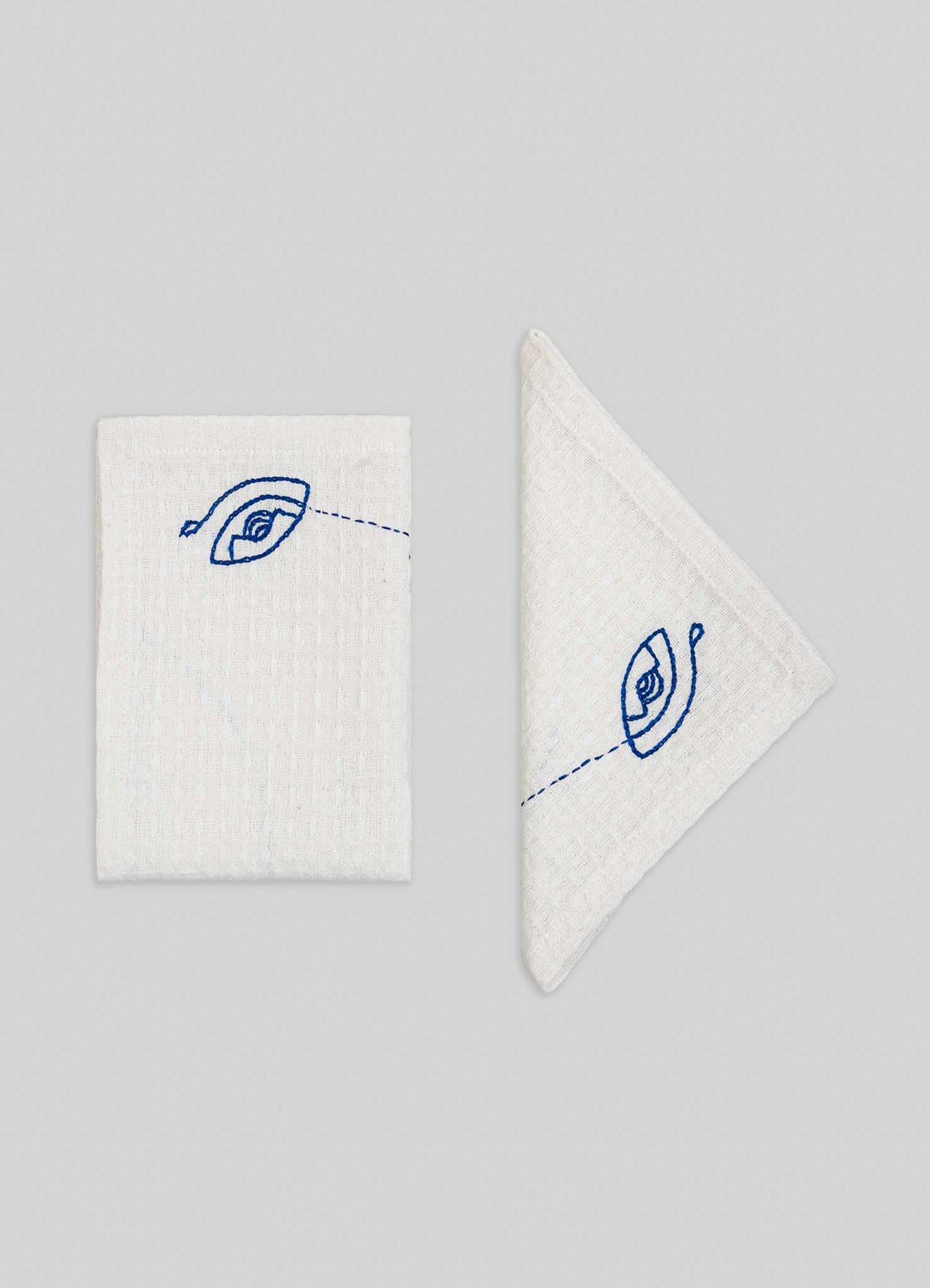 Viya’s Evil Eye Hand Embroidered Ecru Face Towels (set of 4) feature intricate blue Nazar motif designs on white fabric, with one towel folded to display the full eye and another showing part of this protective symbol on a light gray background.
