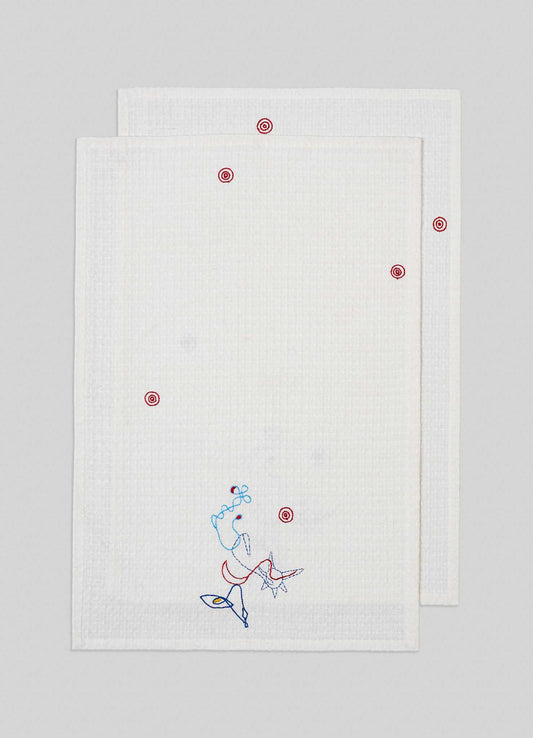 Two Viya Evil Eye Hand Embroidered Ecru Hand Towels (set of 2) are stacked, each with small red circles and an abstract Nazar motif in blue, red, and yellow near the corner. The towels are set against a light gray background.