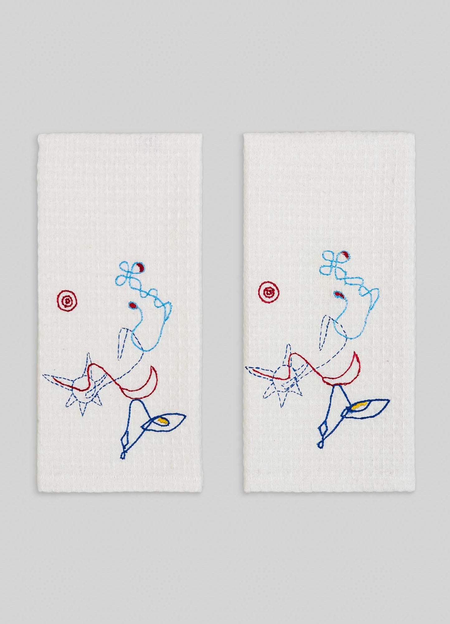 The Evil Eye Hand Embroidered Ecru Hand Towels - Set of 2 by Viya feature colorful abstract embroidery and playful star designs inspired by the Nazar motif, adding a protective totem touch against their light gray background.