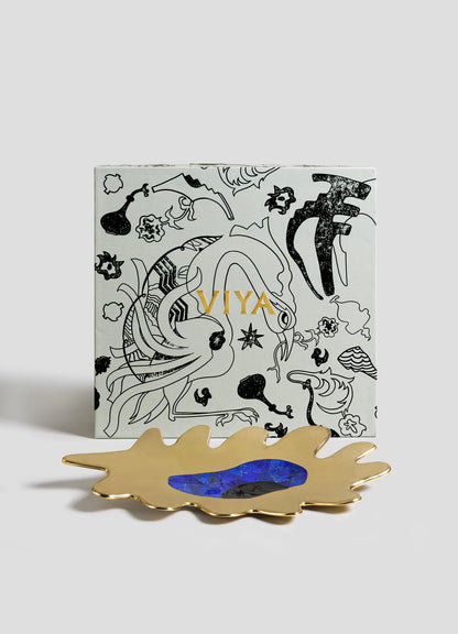 A square VIYA box with gold lettering and black abstract bird-like designs stands behind the Evil Eye Bowl Small, an irregularly shaped gold tray with a blue center, all set against a plain light-gray background.