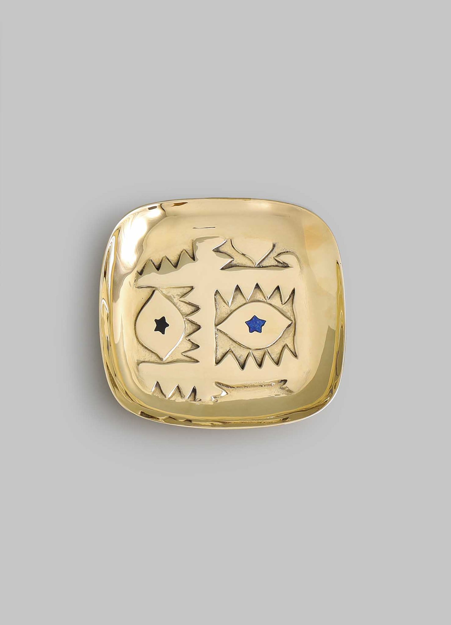 The VIYA Evil Eye Trinket Tray Small is a square gold dish with rounded edges, featuring abstract engraved eyes—one black, one blue with stars—reminiscent of the Nazar amulet for protection, shown on a plain gray background.