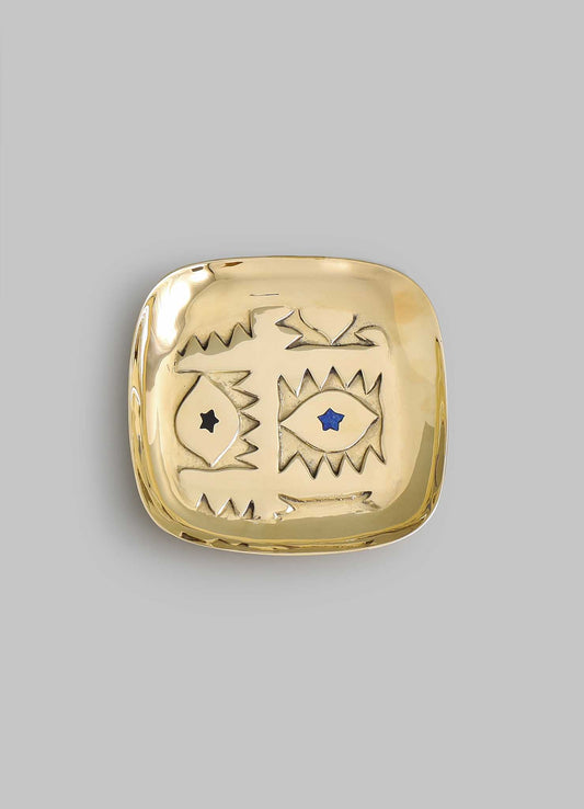 The VIYA Evil Eye Trinket Tray Small is a square gold dish with rounded edges, featuring abstract engraved eyes—one black, one blue with stars—reminiscent of the Nazar amulet for protection, shown on a plain gray background.