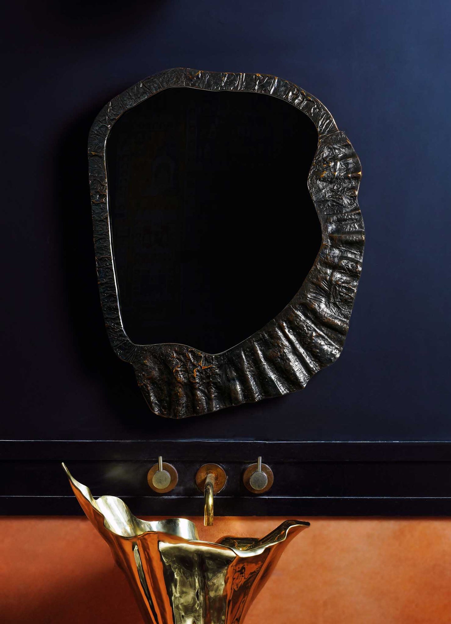 An irregular-shaped mirror with a textured black and gold metallic frame hangs on a dark wall above a brass and black sink with a bent, sculptural design. The overall style appears contemporary with metallic accents and organic forms.