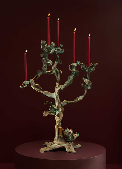 The VIYA Glory Lily Candelabra, inspired by the elegant Glory Lily flower, features sculptural cast brass branches holding five red candles, shown on a round maroon pedestal against a deep maroon background.