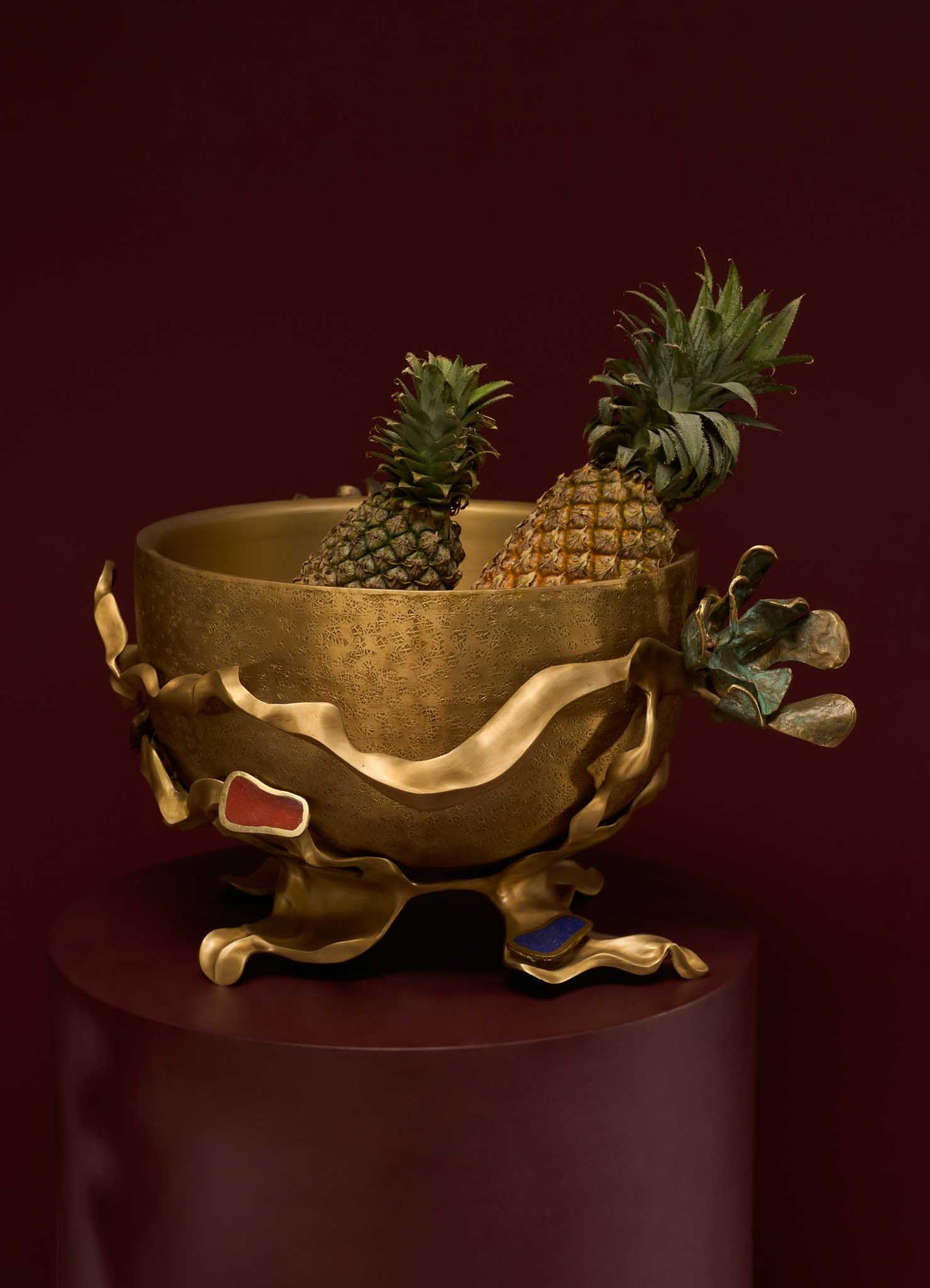 A decorative brass bowl with an antique finish, featuring organic, flowing edges with colored enamel accents, contains two pineapples and is displayed against a dark background.