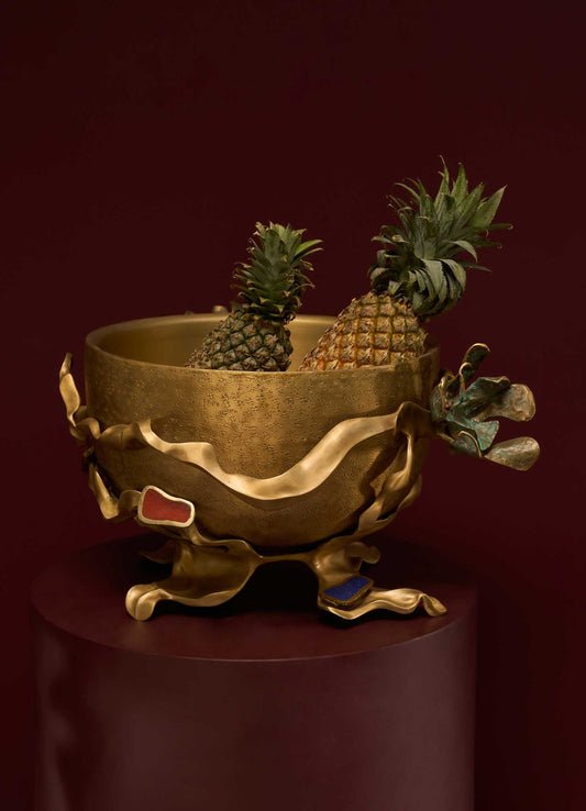 A decorative brass bowl with an antique finish, featuring organic, flowing edges with colored enamel accents, contains two pineapples and is displayed against a dark background.