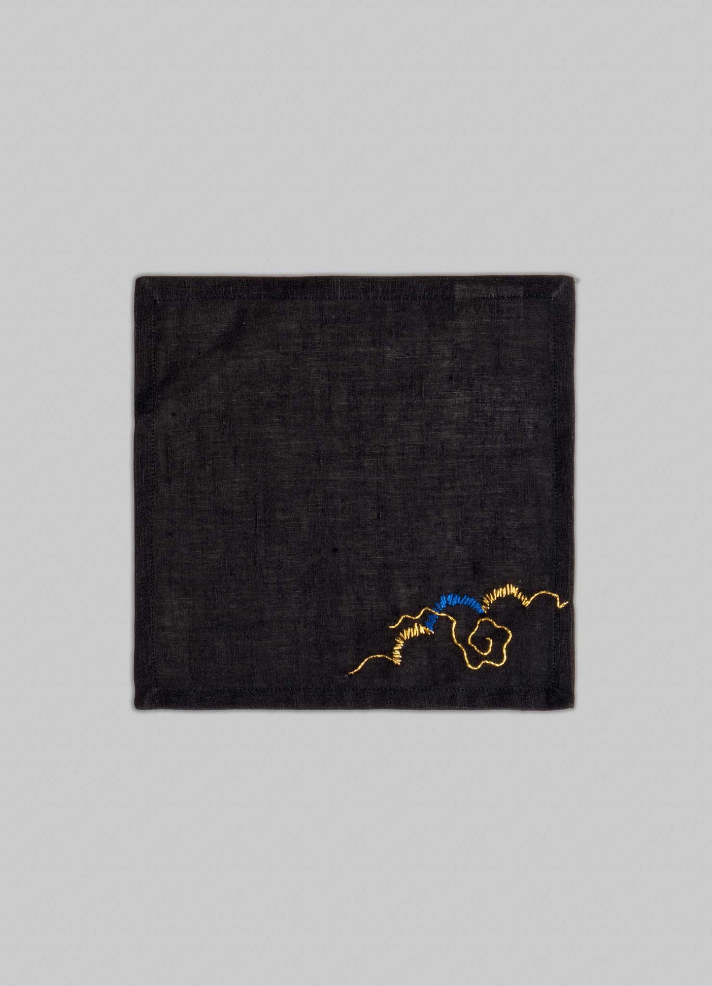 The Viya Gajasimha Hand Embroidered Black & Gold Cocktail Napkins, set of 4, feature small gold and blue floral embroidery in one corner, inspired by the mythical guardian from ancient Indian art. Set against a plain light gray background.