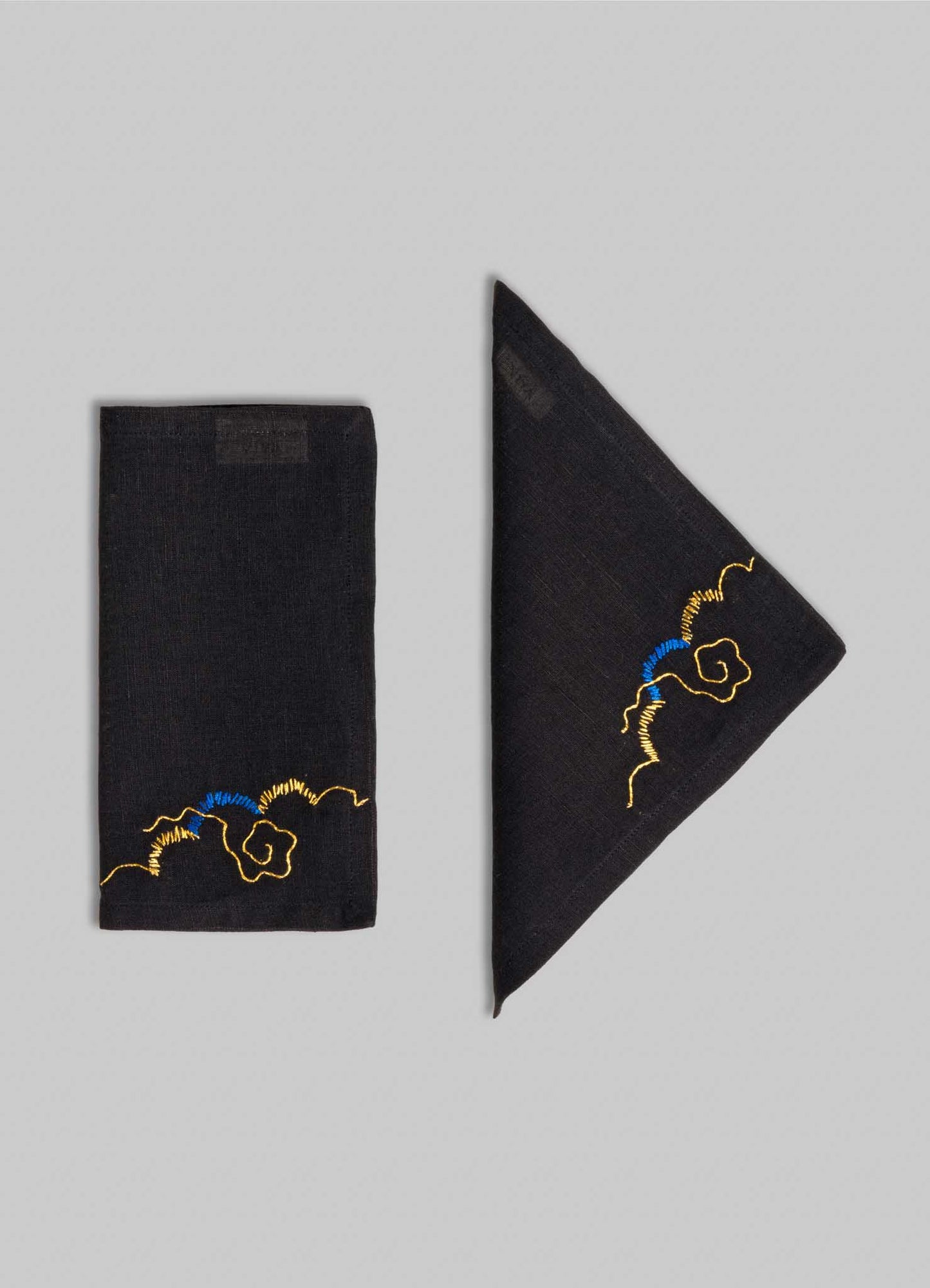Two black Gajasimha Hand Embroidered Black & Gold Cocktail Napkins by Viya, one folded into a rectangle and the other a triangle, showcased on a gray background with gold embroidery inspired by ancient Indian art in one corner.