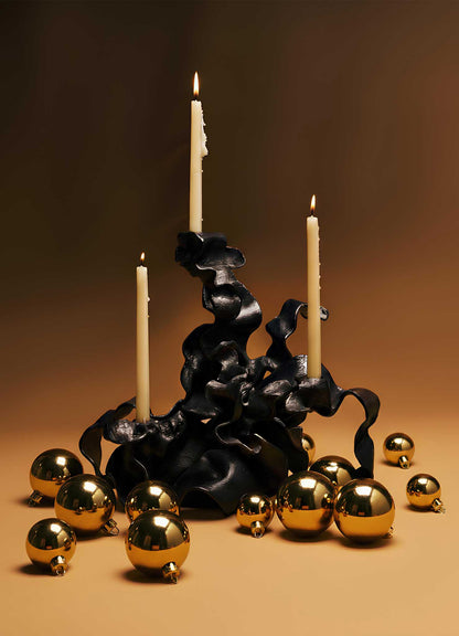 The Hundred Petal Rose Bronze Candelabra by VIYA features three ivory candles in a sculptural black holder, accented with gold ornaments on a warm brown backdrop—two candles lit, one unlit—for elegant home decor.