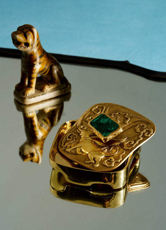 The VIYA Leophant Trinket Box, a small golden box with an ornate design and green gem, sits on a mirror with its reflection. Behind it, a tiger figurine evokes the mythical guardian spirit seen in Hindu Buddhist art, also mirrored on the surface.