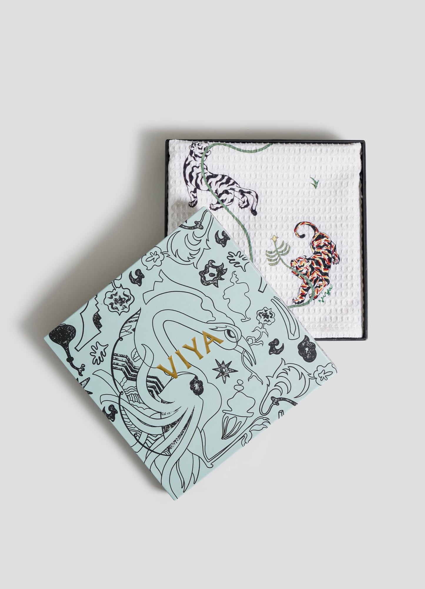 A decorative Viya box with gold "VIVA" lettering is partially open, revealing a set of two Lounging Tiger Hand Towels featuring colorful, embroidered animals inspired by the Indian menagerie.