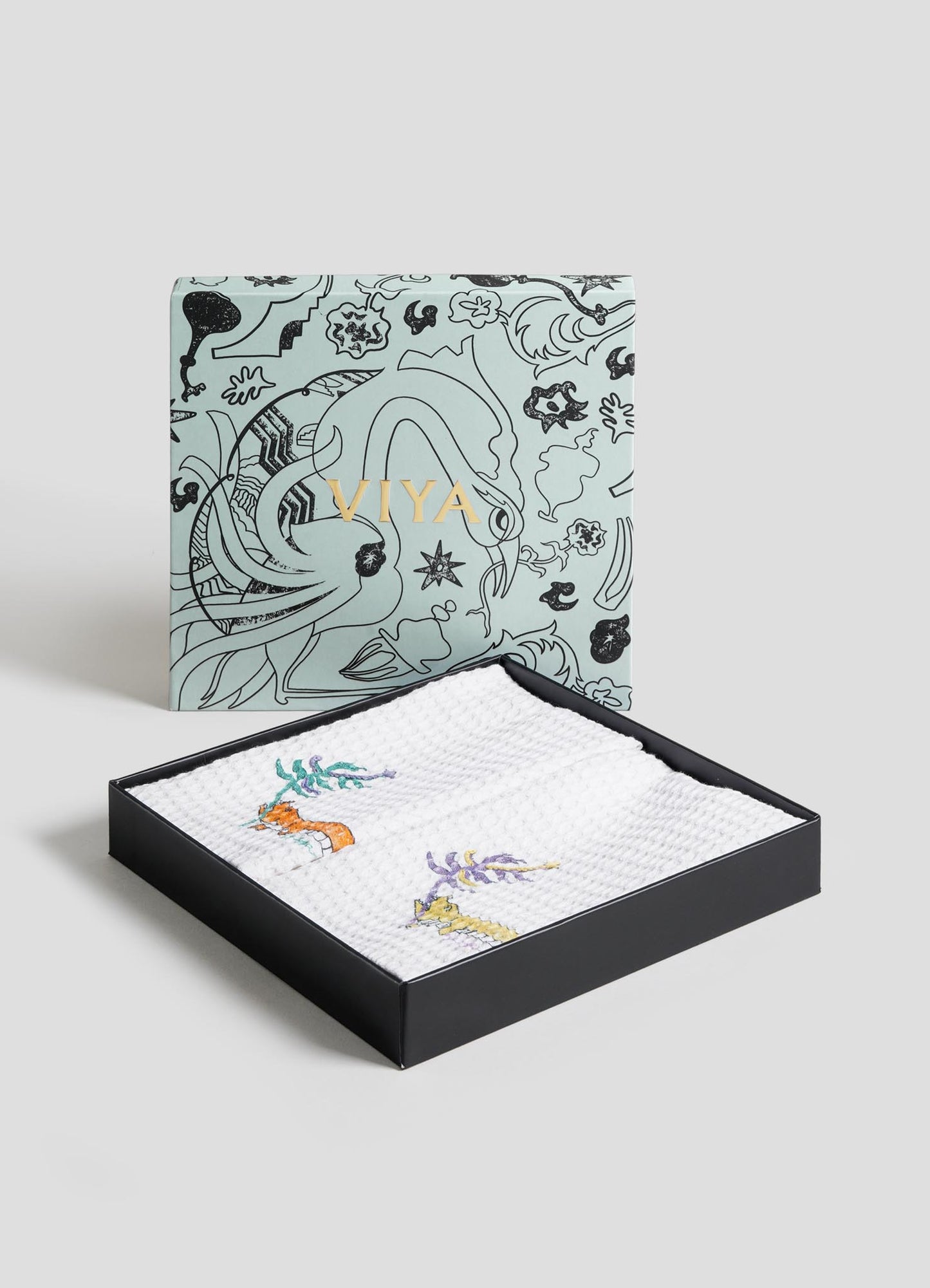 The Majestic Blackbuck Hand Towels - Set of 2 by Viya, featuring Indian wildlife-inspired floral patterns, are neatly stacked on a black metal tray against a plain light blue background, with one towel draped over the tray’s edge.