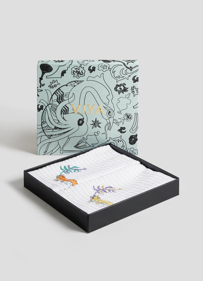 The Majestic Blackbuck Hand Towels - Set of 2 by Viya, featuring Indian wildlife-inspired floral patterns, are neatly stacked on a black metal tray against a plain light blue background, with one towel draped over the tray’s edge.