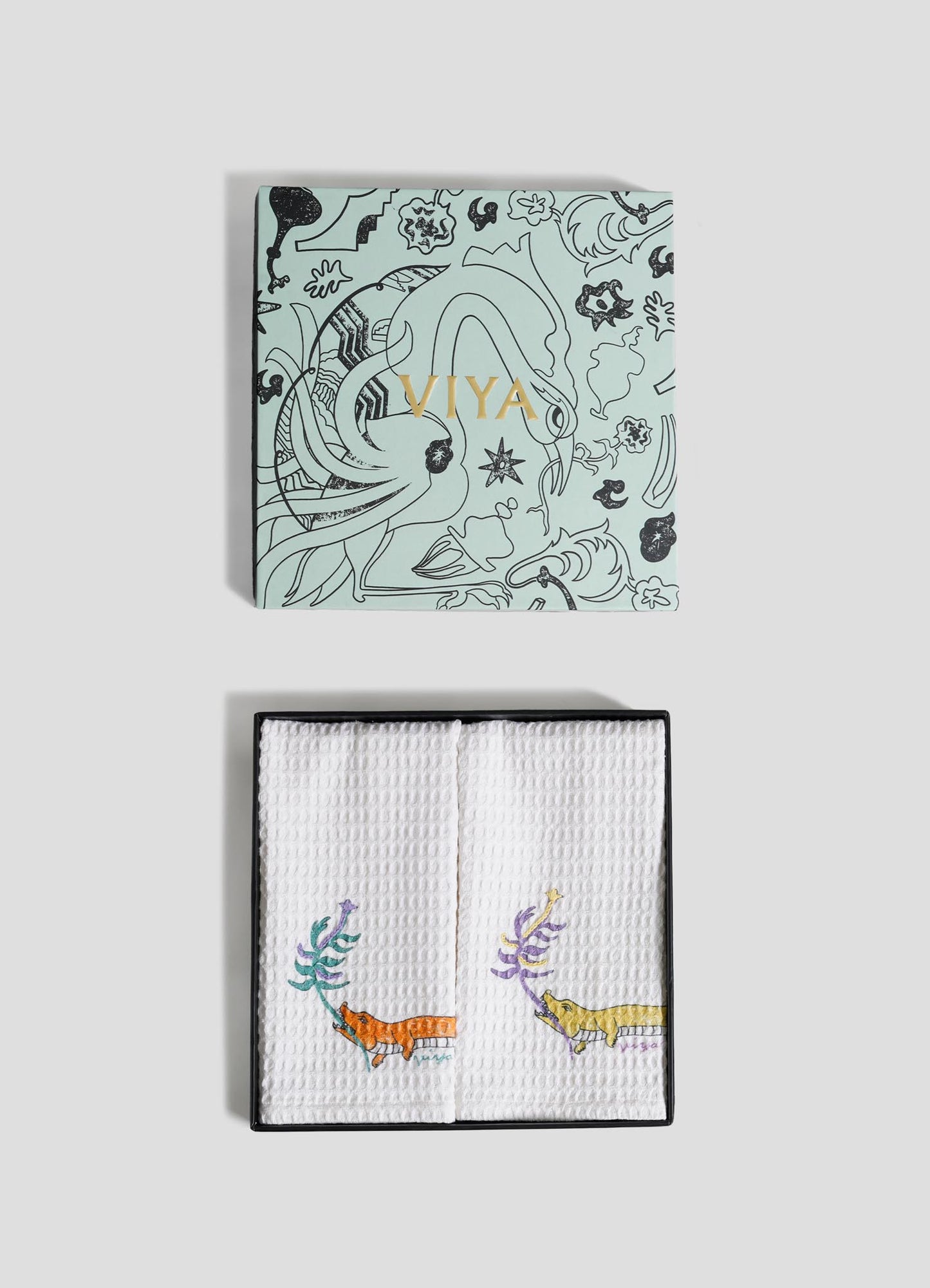 The Lurking Crocodile Hand Towels by Viya come as a set of 2, packaged in a decorative black box with "Viya" in gold, featuring folded white towels embroidered with vibrant aquatic animals and plants.