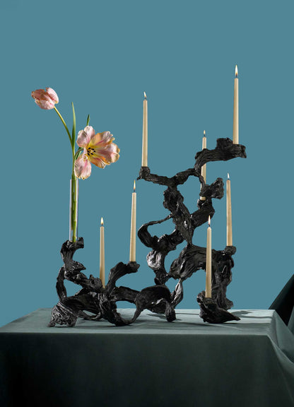 The VIYA Leher Candelabra Large, with a twisted organic black metal design, holds six taper candles and two pink flowers on a teal-draped table set against a matching teal background.