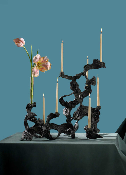 The VIYA Leher Candelabra Large, with a twisted organic black metal design, holds six taper candles and two pink flowers on a teal-draped table set against a matching teal background.