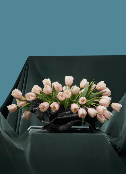 A floral arrangement of pale pink tulips in the VIYA Leher Vessel sits on a table draped with dark green Leheriya textile, set against a solid teal background.