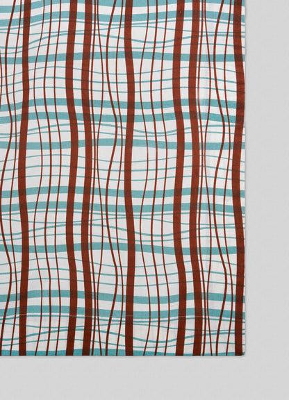 The Viya Madras Check Aqua & Rust Table Cloth features a rectangular design with wavy brown, teal, and red lines forming a modern take on classic Indian Madras checks, displayed diagonally on a light gray surface.