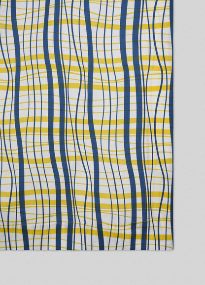 The Viya Madras Check Blue & Yellow Table Cloth features a wavy, irregular plaid of blue and yellow lines on white, shown on a light gray surface.