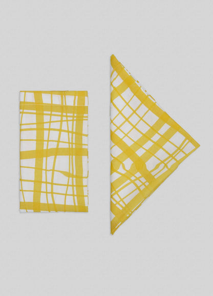 Viya’s Madras Check Yellow Napkins – Set of 4 are displayed on a light gray background, with two white napkins featuring yellow Madras checks folded into rectangle and triangle shapes, highlighting contemporary patterns from the Indian subcontinent.