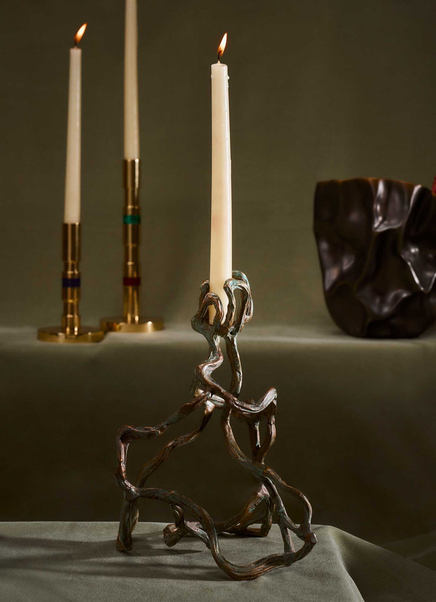 A sculptural candle holder made of twisted, metallic material, supporting a tall white taper candle, with a dark, irregularly shaped ceramic or resin vessel nearby. In the background, there are two additional candlesticks with white candles and gold accents.