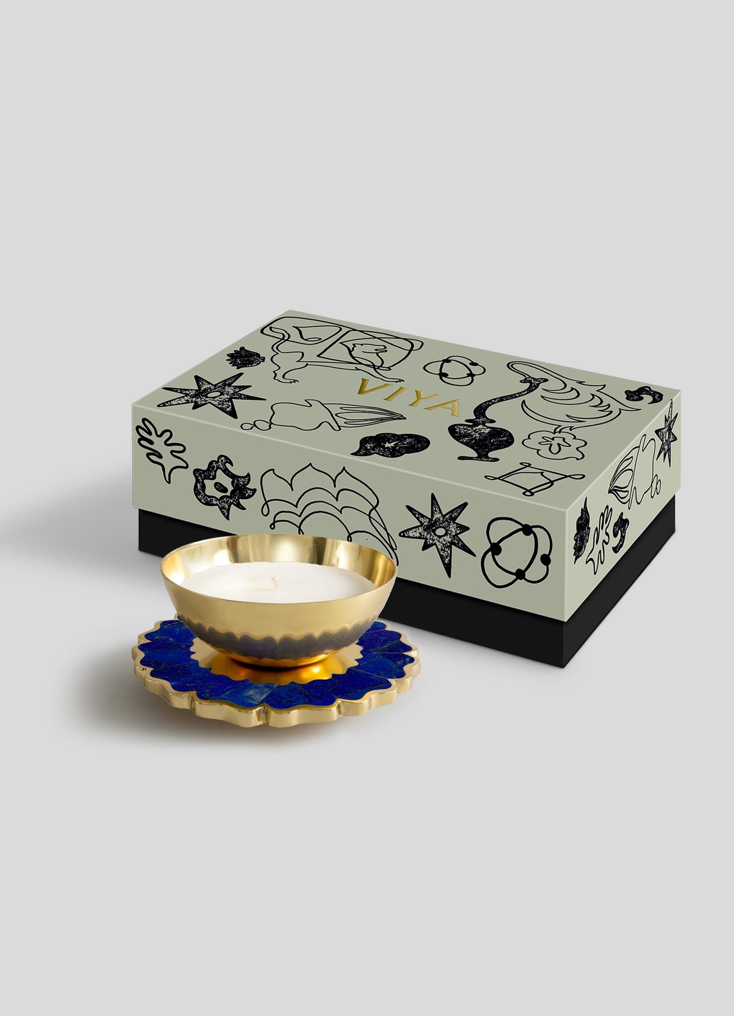 The Viya Mystical Marigold Blue - Scented Candle Diya Large, in a gold bowl, rests on a blue flower-shaped coaster before a light gray gift box with black and gold designs.