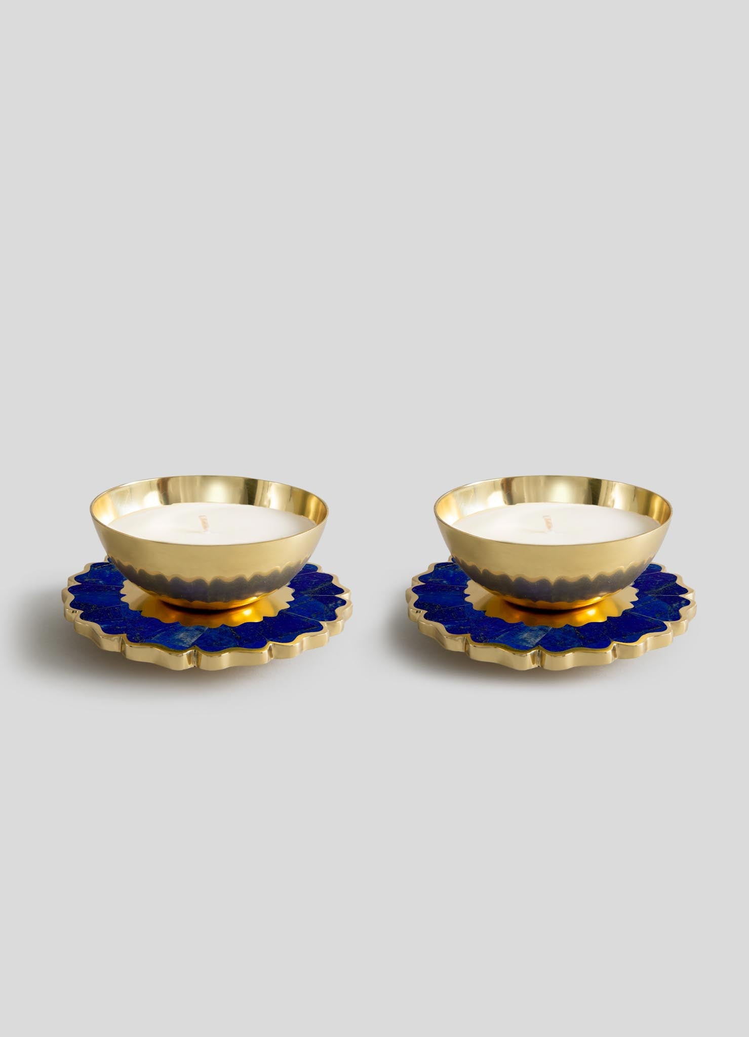 The Viya Mystical Marigold Blue Scented Candle Diya Small Set of 2, featuring gold metal holders with scalloped edges, is displayed on blue and gold floral coasters against a light gray background.