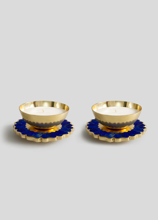 The Viya Mystical Marigold Blue Scented Candle Diya Small Set of 2, featuring gold metal holders with scalloped edges, is displayed on blue and gold floral coasters against a light gray background.