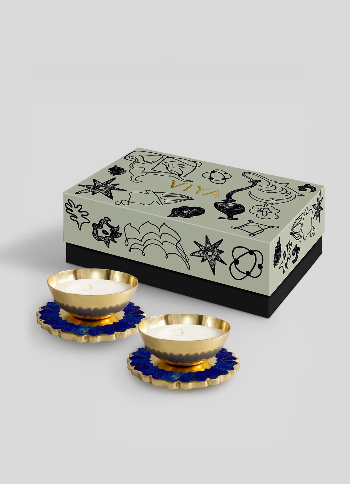 The Viya Mystical Marigold Blue Scented Candle Diya Small Set of 2, with blue and gold floral bases, pairs beautifully with a gray-lidded decorative box featuring black designs crafted using ancient metalworking techniques.