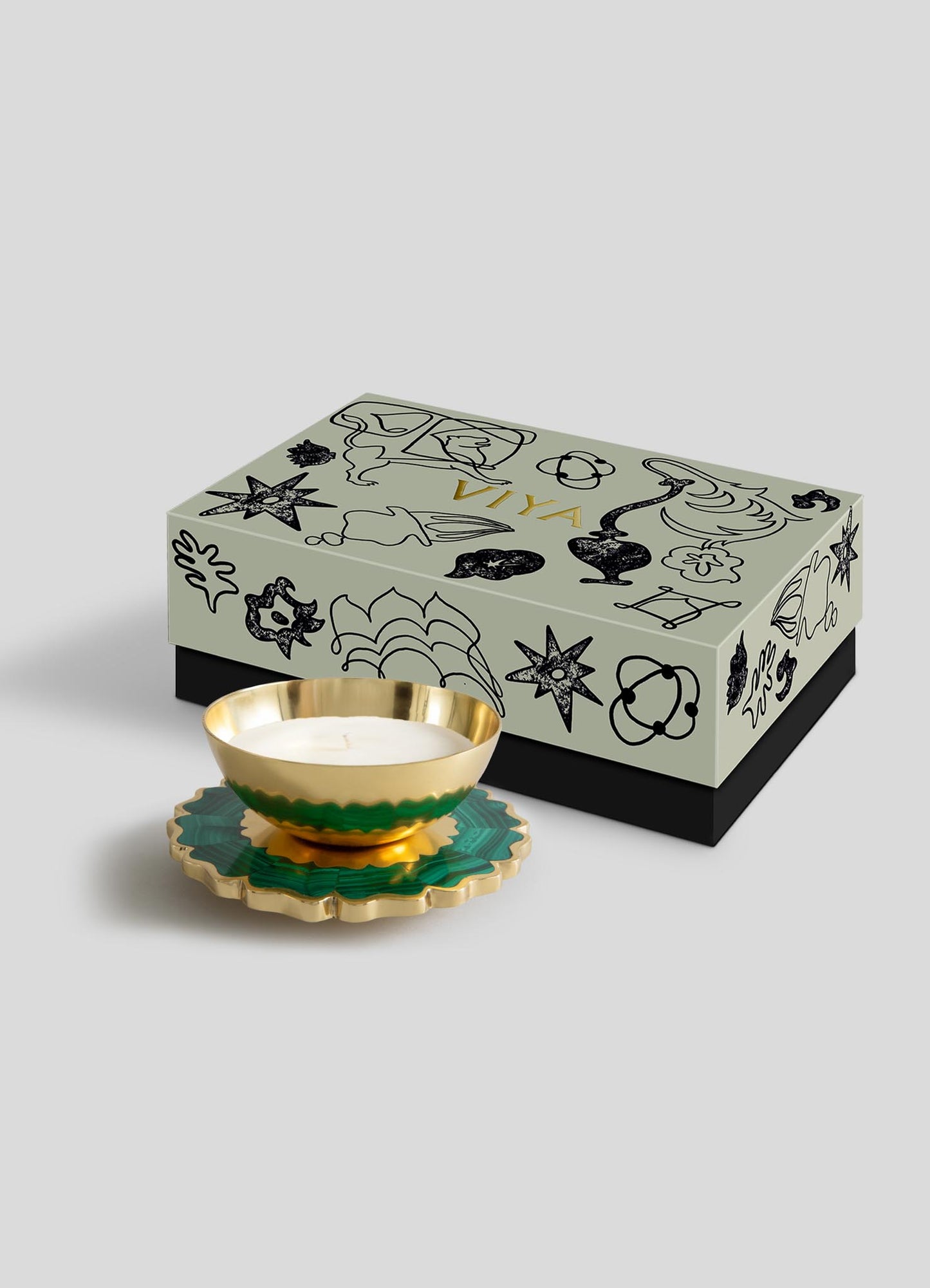 The Mystical Marigold Green - Scented Candle Diya Large by Viya, in a gold and green holder crafted with ancient techniques, sits before a light grey VIVA box with black illustrations, adding heirloom charm to modern decor.