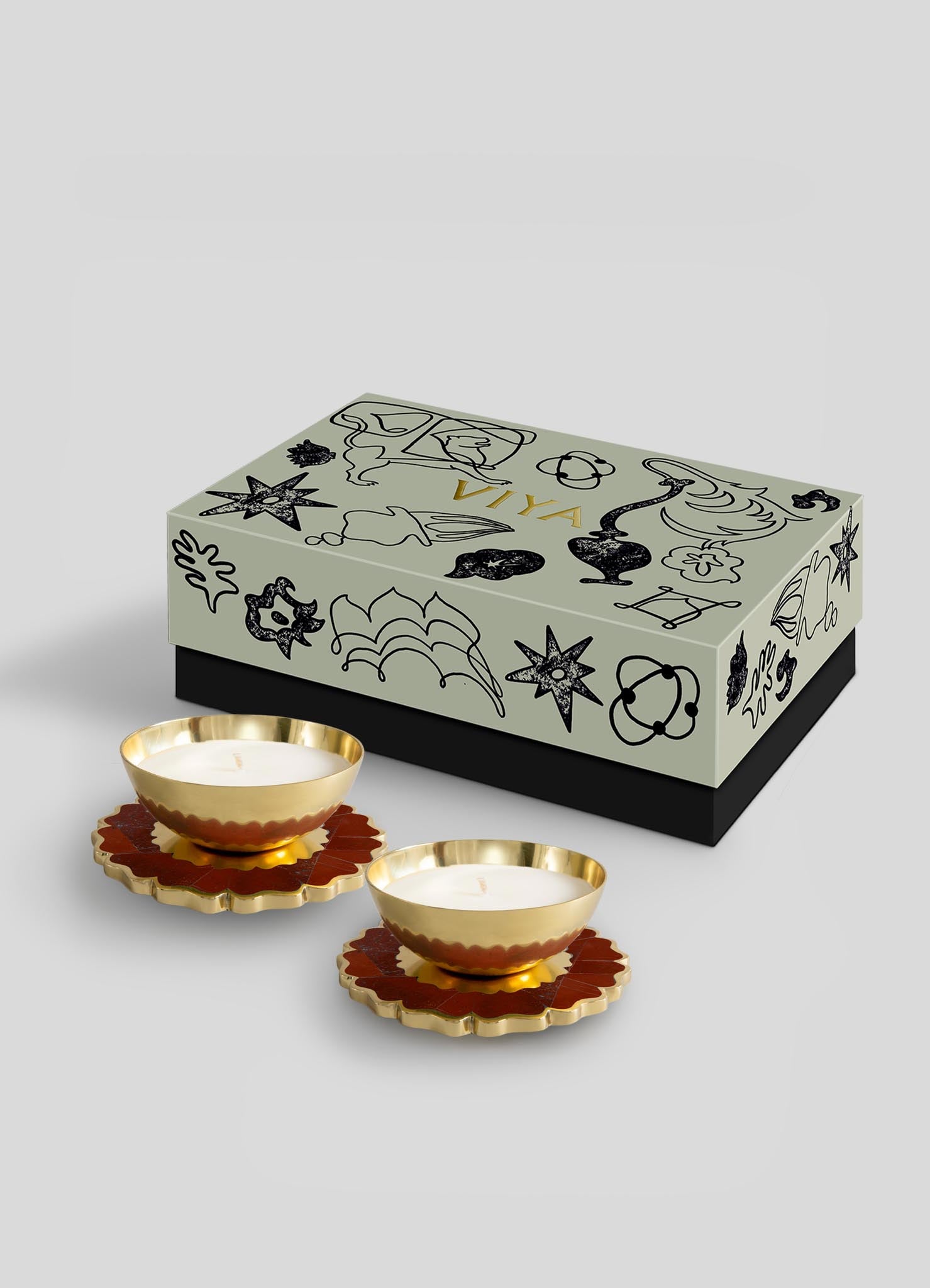 The Mystical Marigold Red Scented Candle Diya Small Set of 2 by Viya showcases intricate black line art, gold-rimmed holders crafted with ancient techniques, and brown flower-shaped coasters—all set against a plain gray background.