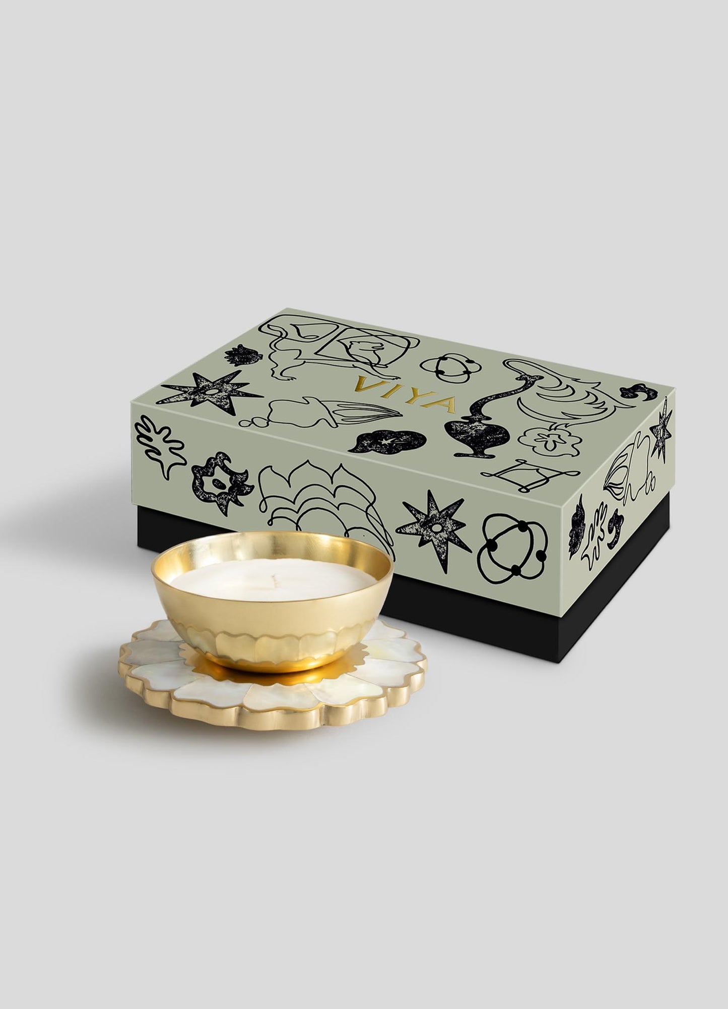 The Viya Mystical Marigold White - Scented Candle Diya Large features a gold bowl inspired by ancient metalwork, resting on a scalloped white and gold base, beside a light gray box with Pichwai-style black sketches.