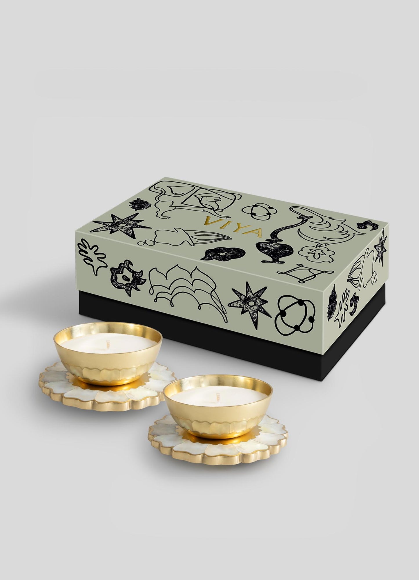 The Viya Mystical Marigold White Scented Candle Diya Small Set of 2 features gold and white candles on flower-shaped bases, displayed before a black and gold Indian-inspired decorative box against a light gray background.