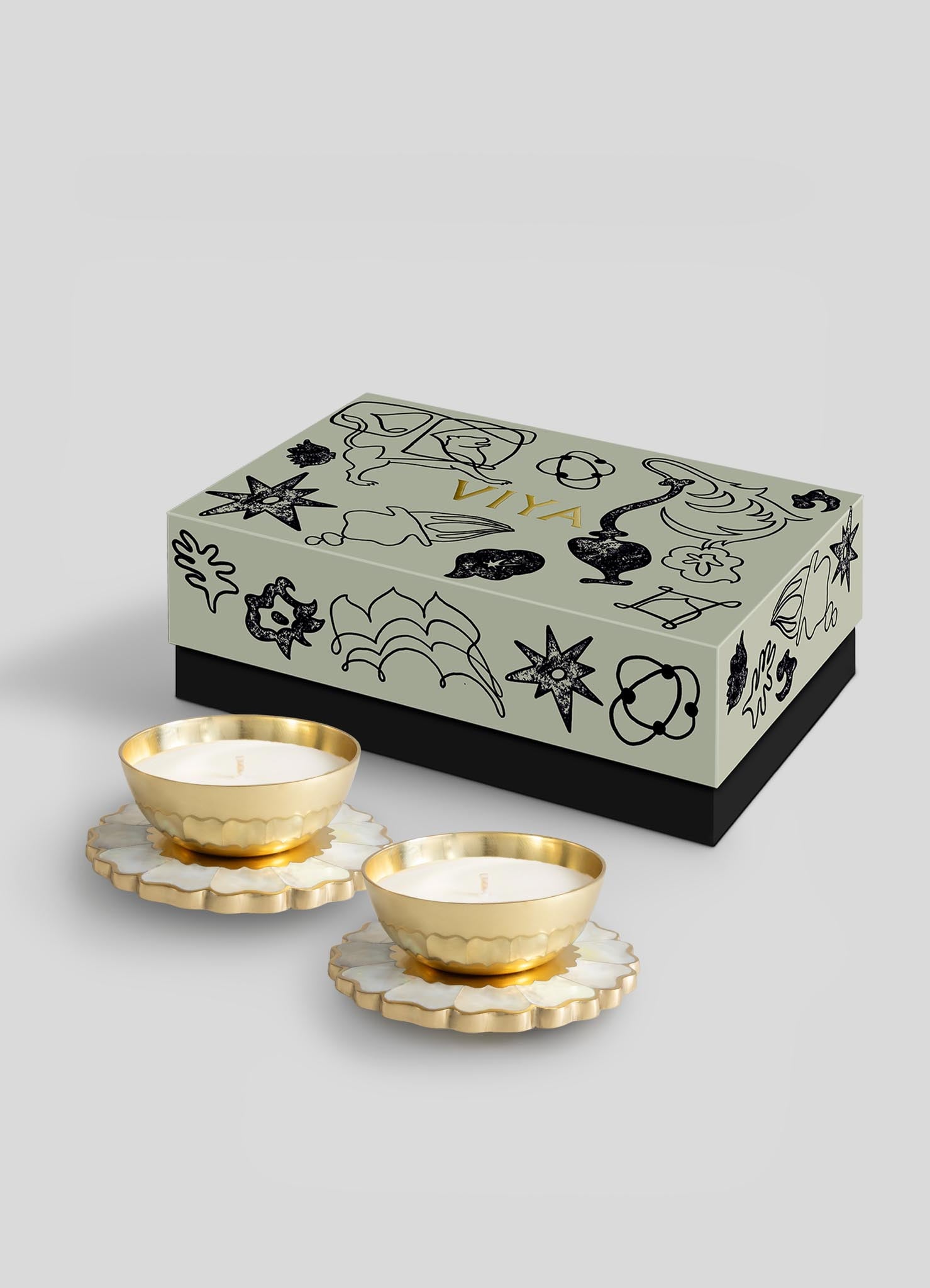 The Viya Mystical Marigold White Scented Candle Diya Small Set of 2 features gold and white candles on flower-shaped bases, displayed before a black and gold Indian-inspired decorative box against a light gray background.
