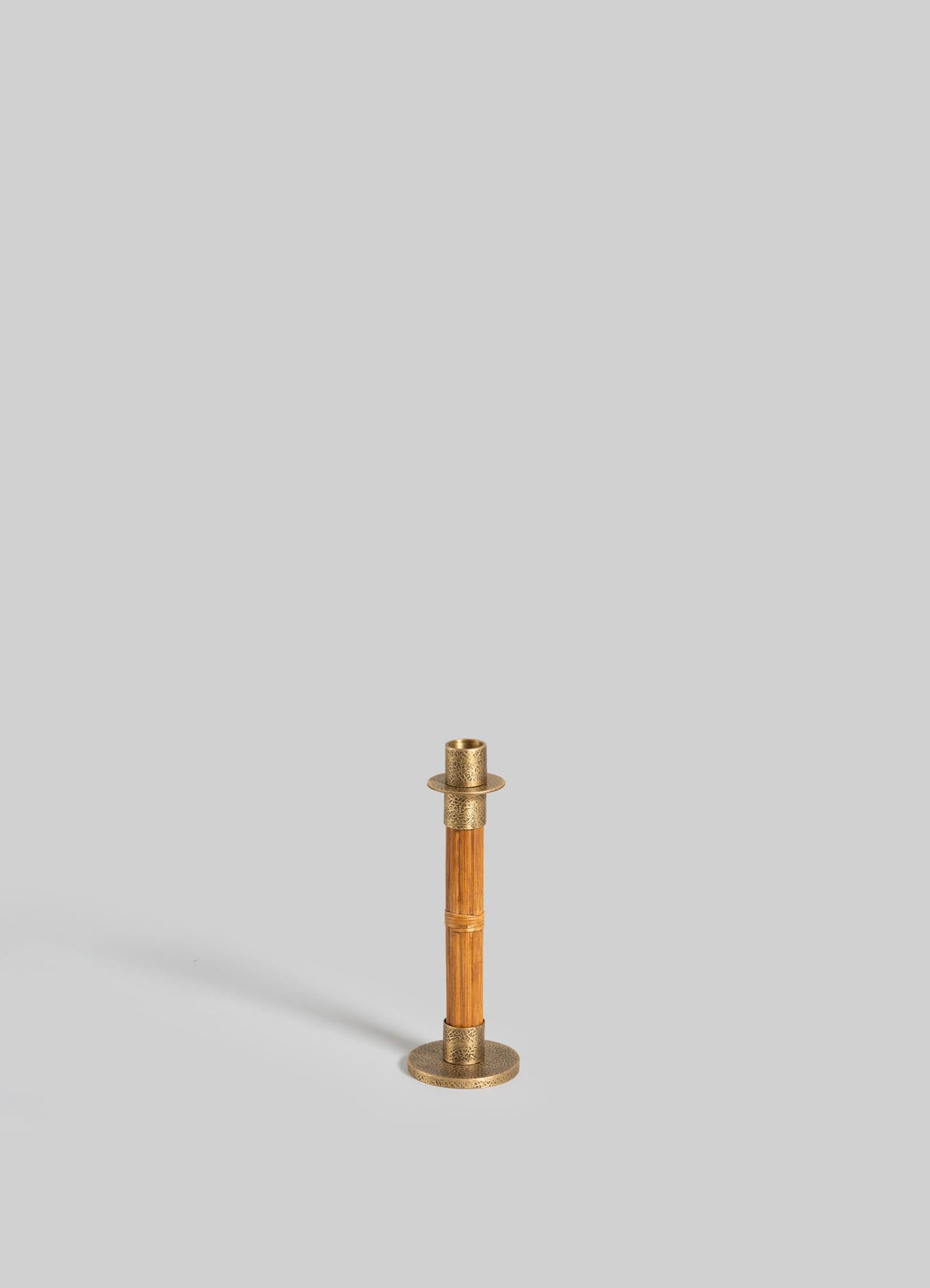 The VIYA Namsai Candle Holder Small features a brass finish with a cylindrical stem and round base, standing upright on a plain light gray background and casting a subtle shadow to the left.