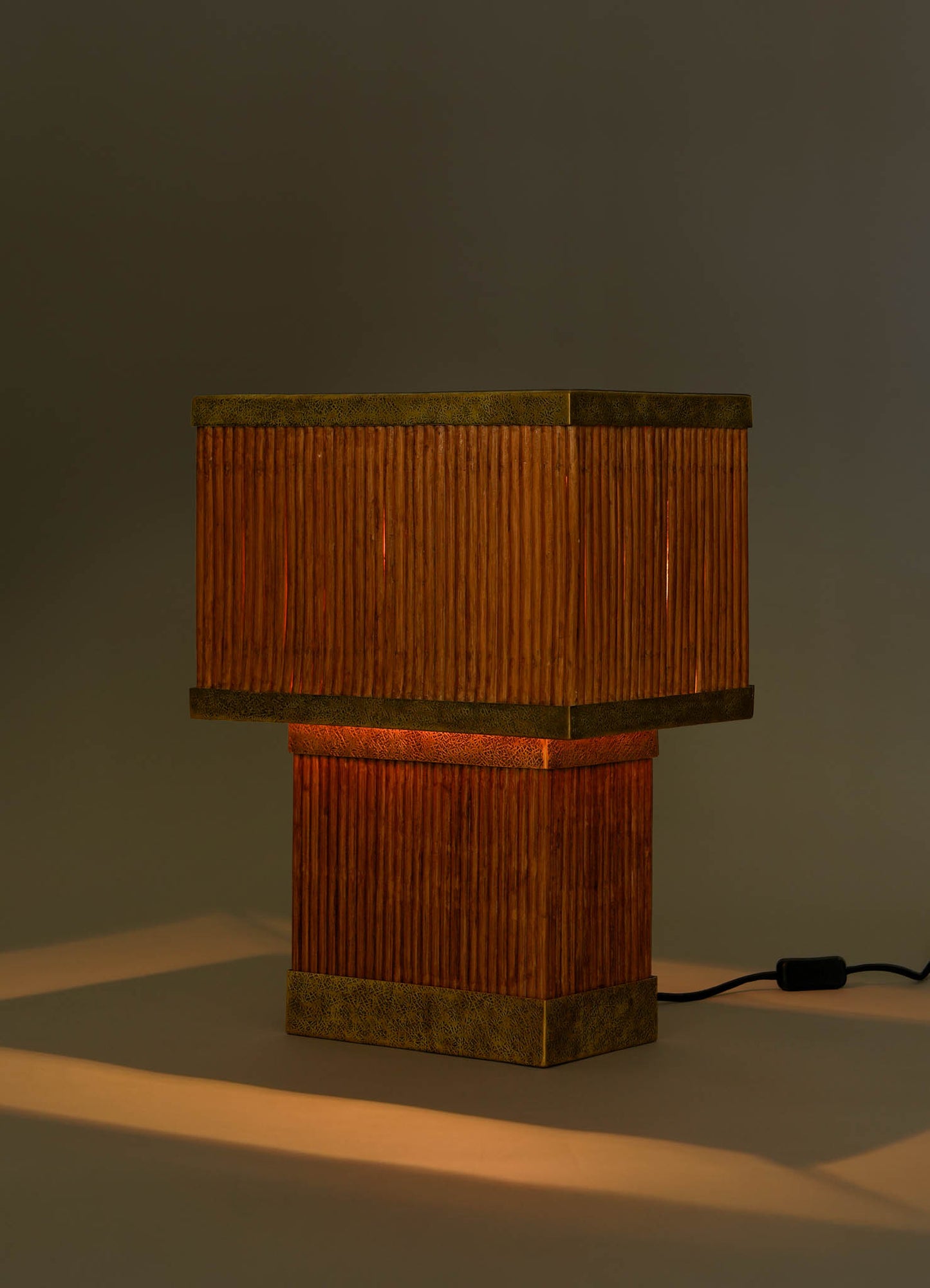 The VIYA Namsai Table Lamp Large features a square design with a ribbed wooden base, matching shade, and brass accents. When plugged in, it emits a warm orange glow and casts soft light and gentle shadows on a neutral backdrop.