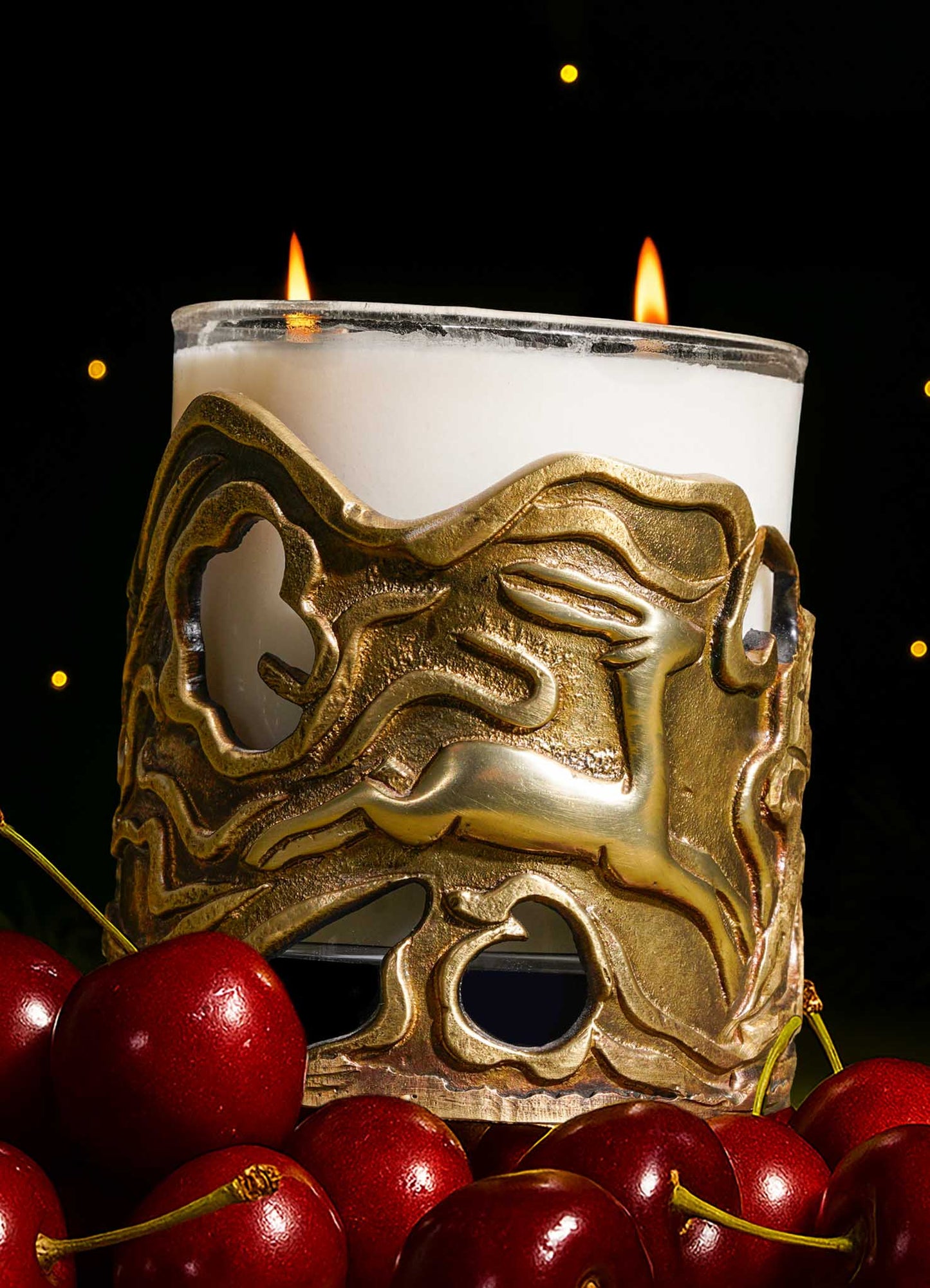 A lit VIYA Patchouli Bloom Candle in a glass holder with an ornate gold animal-shaped cover sits among fresh red cherries, set against a dark background with glowing lights, evoking the essence of Indian fragrances.
