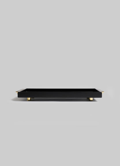The Plaza Vanity Tray by VIYA is a rectangular black tray with gold feet and corner accents, channeling Art Deco glamour. Styled on a light gray background.
