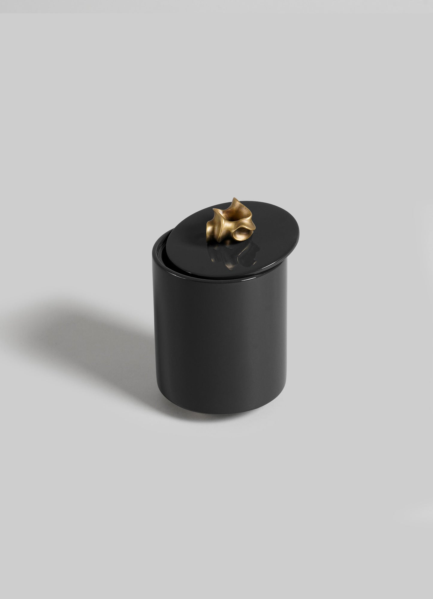 The VIYA Saffron Black Round Container is a sleek, black cylinder with an oval lid and sculptural gold accent inspired by saffron threads, shown against a plain light gray background.