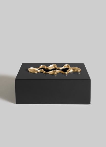 The VIYA Saffron Black Tissue Box Large features a rectangular black design with a sculptural wavy gold accent on the lid, displayed against a plain light gray background.