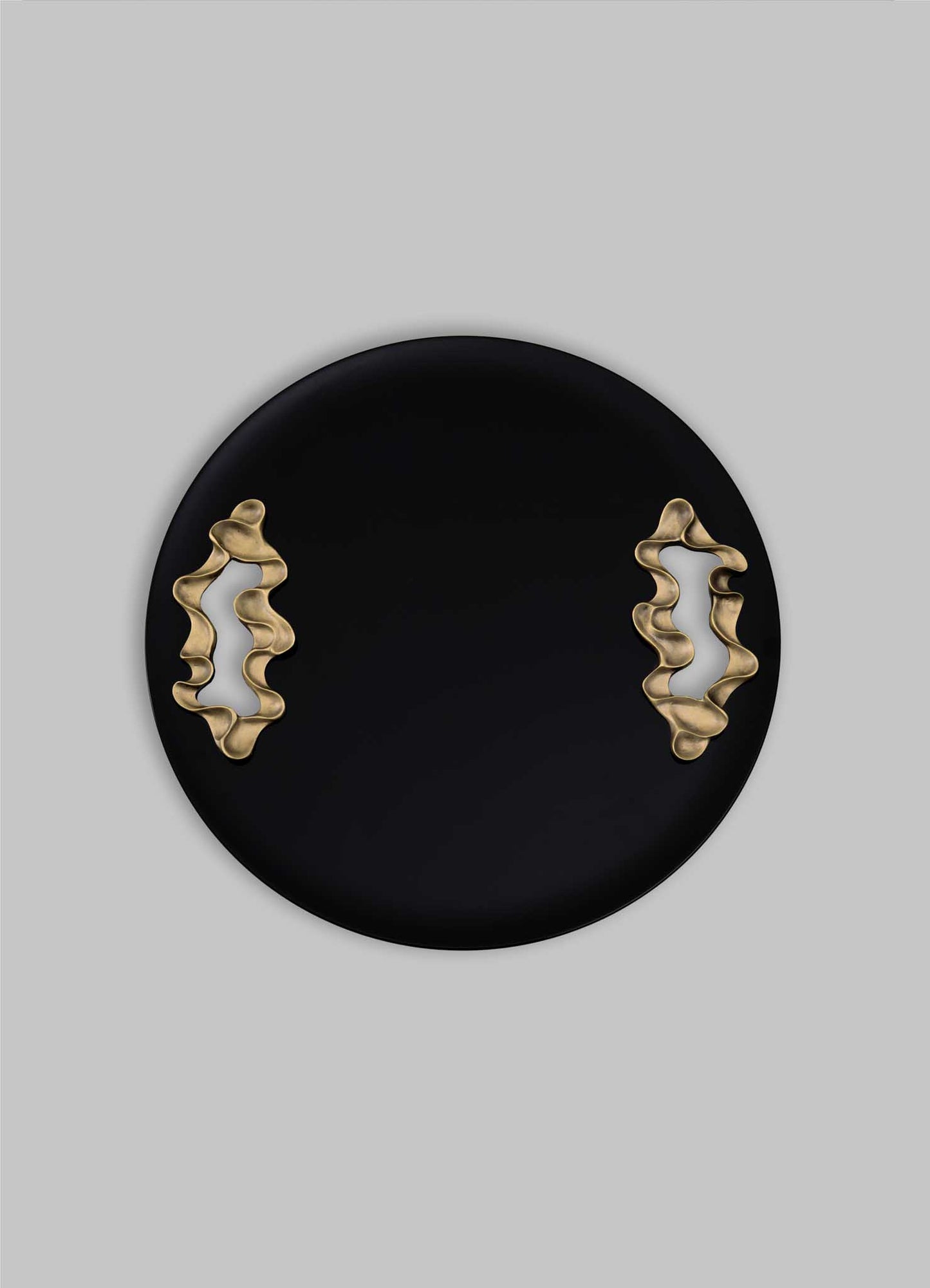 The Saffron Black Tray Large by VIYA features a round black design with two wavy gold accents on opposite sides, reminiscent of contemporary bronzed objects, displayed against a plain gray background.