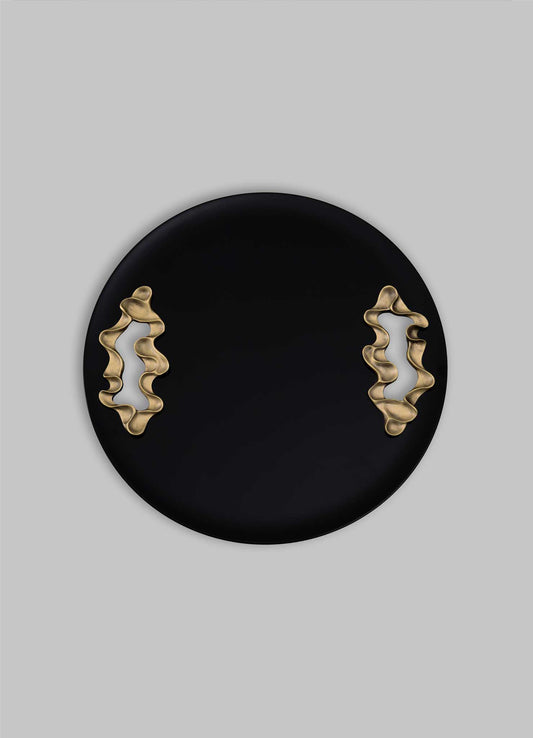 The Saffron Black Tray Large by VIYA features a round black design with two wavy gold accents on opposite sides, reminiscent of contemporary bronzed objects, displayed against a plain gray background.