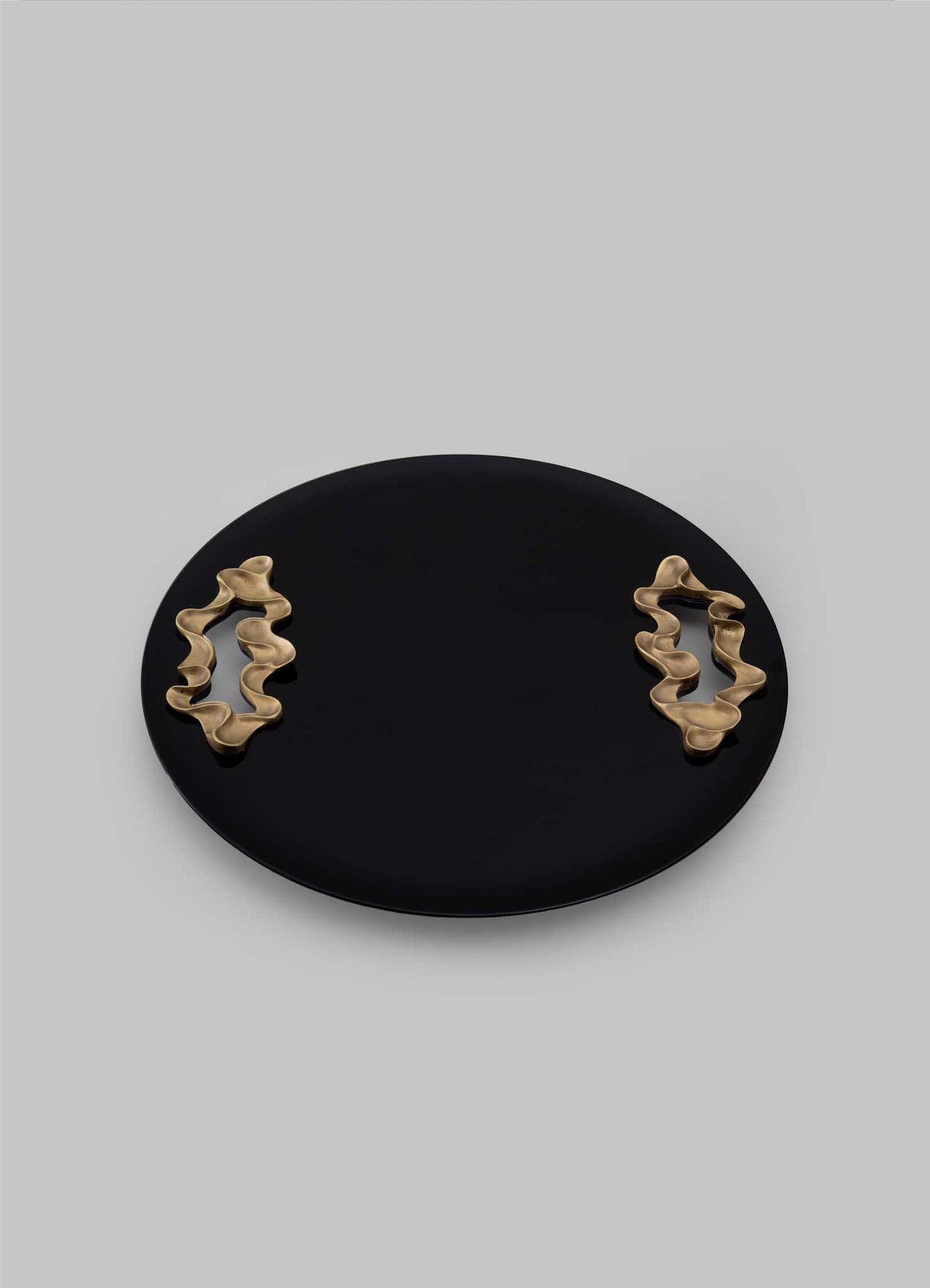 The VIYA Saffron Black Tray Large features a round, flat black design with two abstract wavy gold handles on opposite sides, evoking contemporary elegance against a plain light gray background.