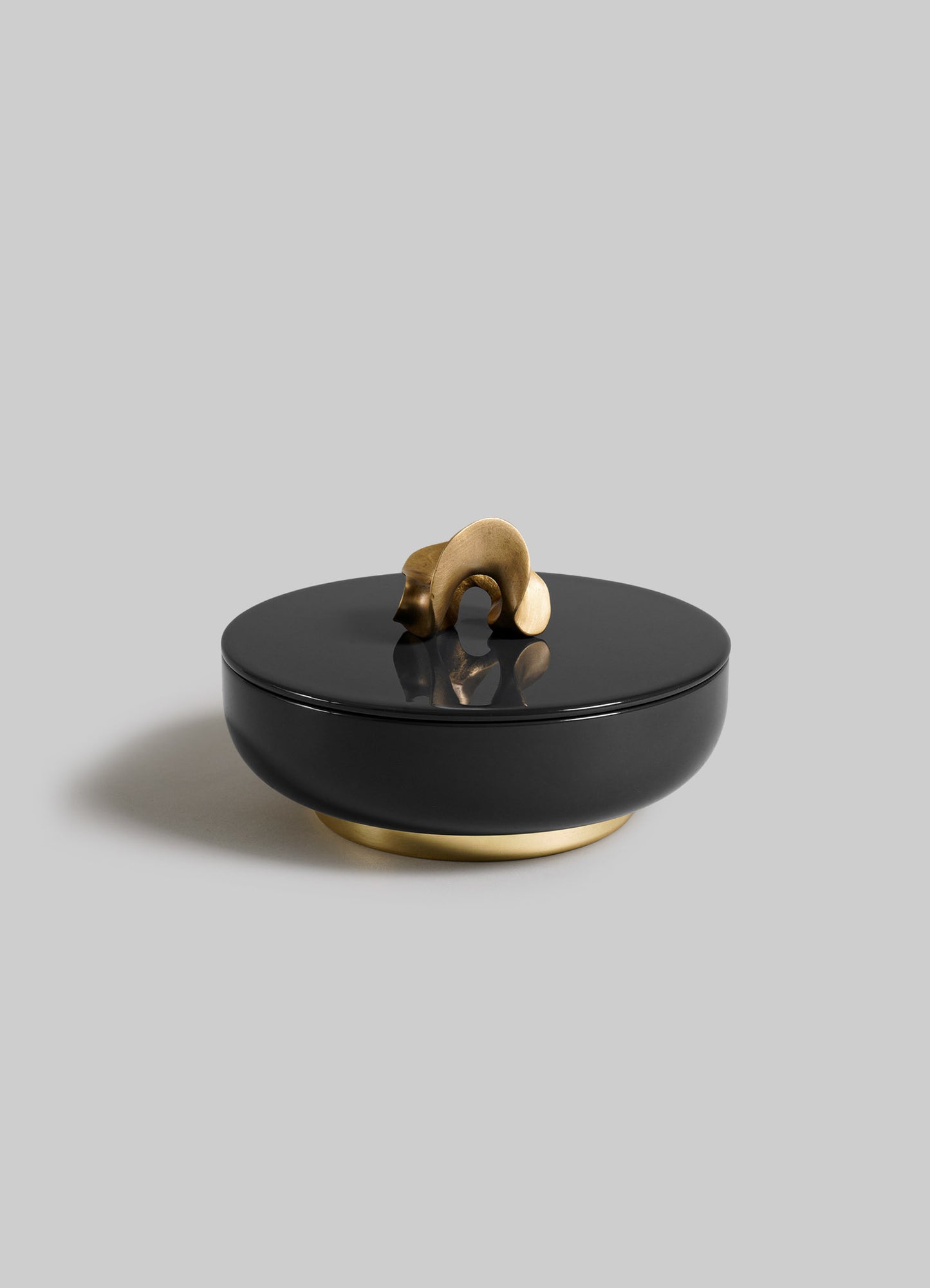 The VIYA Saffron Black Trinket Box Large is a round black container with a lid, featuring an abstract gold handle and matching gold base, elegantly set against a plain light gray background.