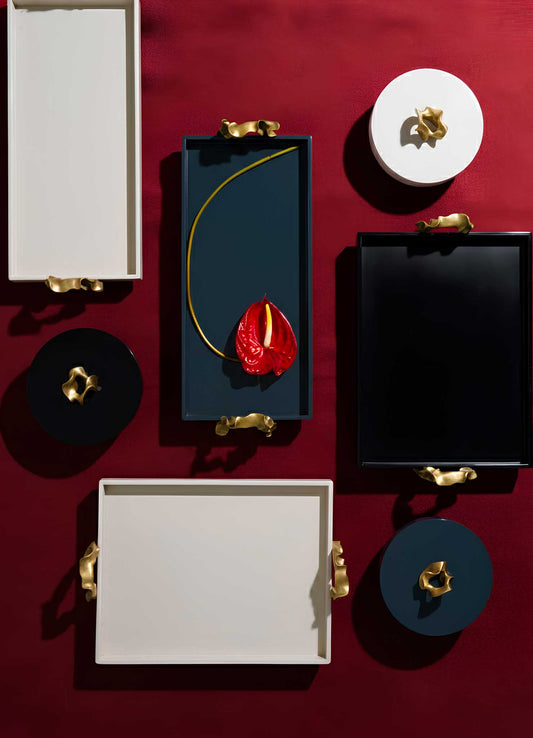 The VIYA Saffron Black Towel Tray is arranged on a deep red surface, holding a single red anthurium flower with a long yellow stem. Gold handles and contemporary bronzed accents highlight its modern rectangular design.