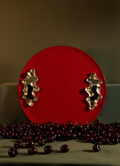The VIYA Saffron Red Tray Small, featuring two wavy gold handles and a round red design, stands upright behind fresh cherries on a green surface with a muted green background.